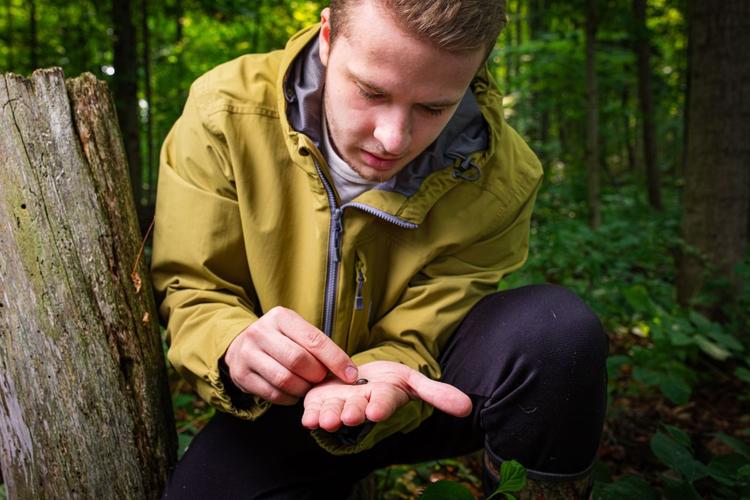 U of G student holds inspecting a tiny specimen in his hand while surrounded by trees.