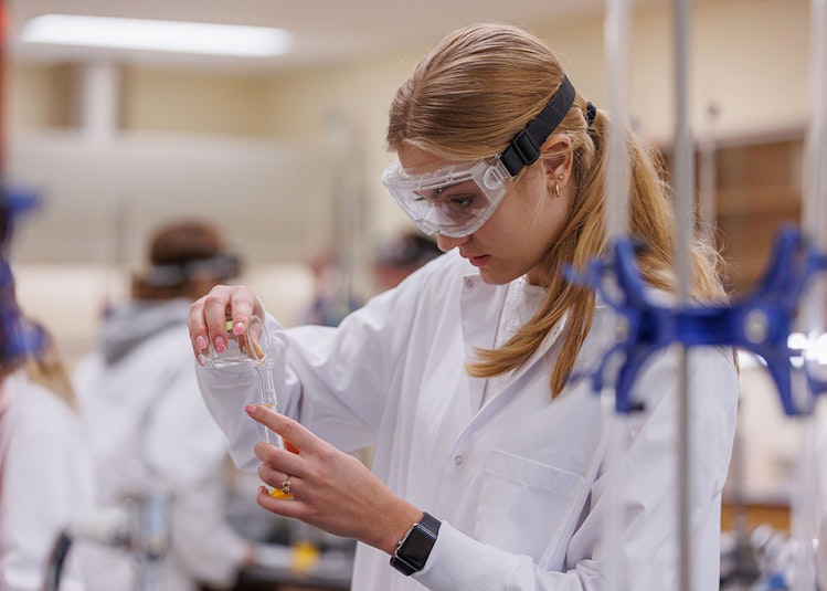 A student works in a chemistry lab, pouring someone from one beaker to another while wearing PPE