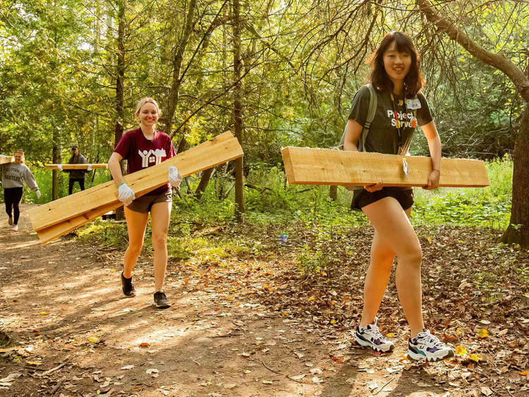 two people carrying wood planks along forested trail