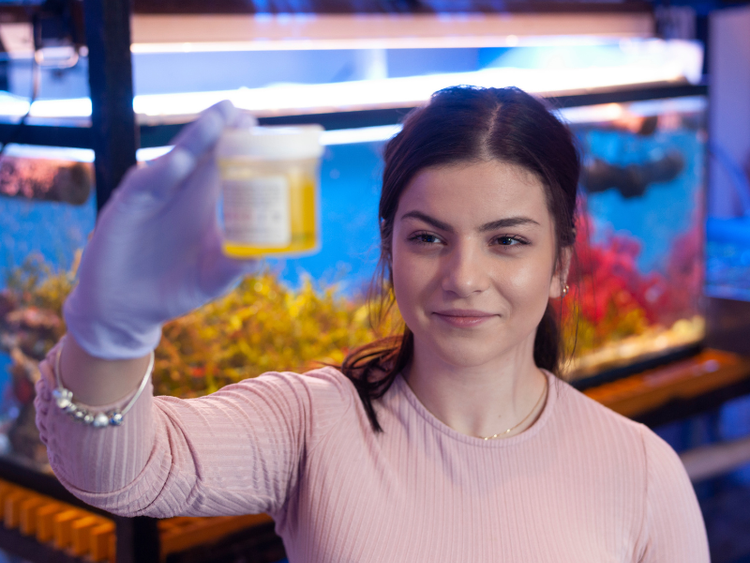 A student stands in front of an aquarium and holds up a container with liquid inside.