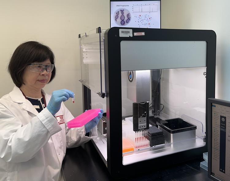 Shu Chen in a lab coat working with samples in a pink rack next to an open PCR (Polymerase Chain Reaction) machine, with a monitor displaying samples in the background.