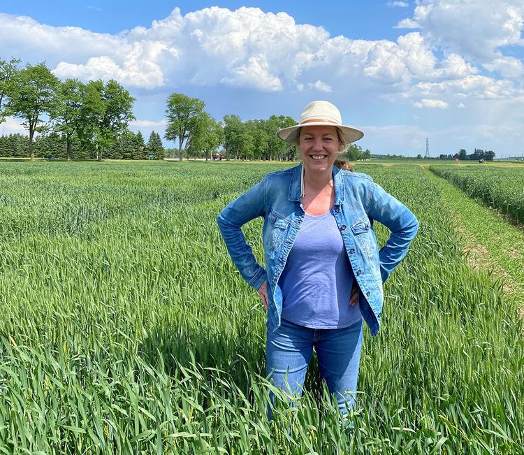 Dr. Helen Booker in a straw hat and denim jacket standing happily in a lush green wheat field under a clear blue sky.