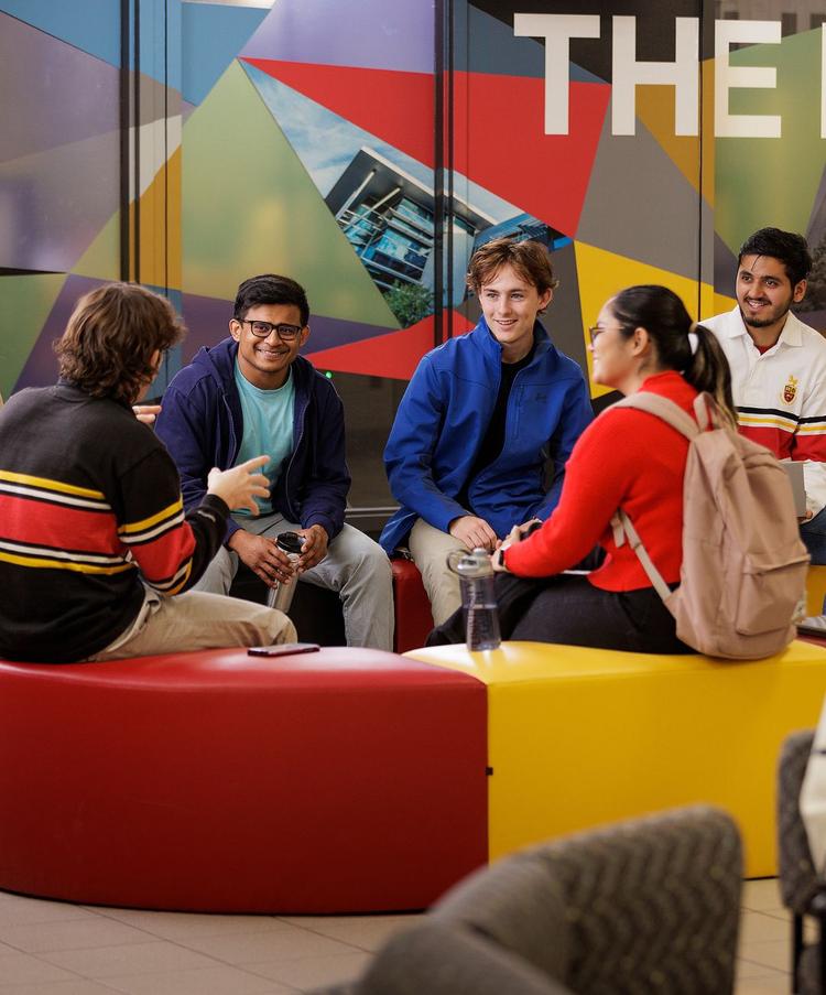 Group of university students sitting together in a colorful campus common area, talking and studying.