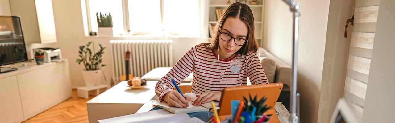 Female sitting at a desk, writing in notebook while looking at the tablet screen in front of her