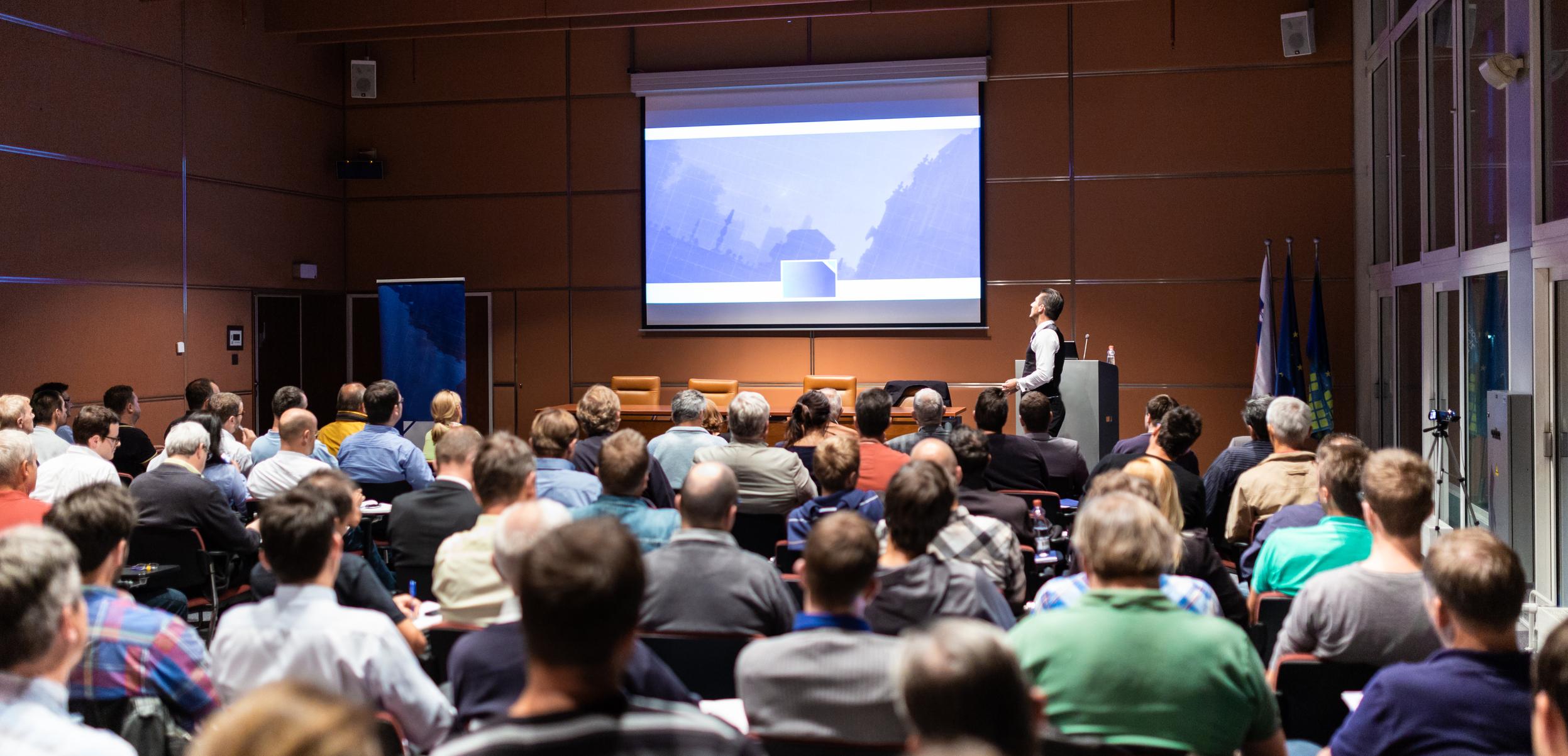 A speaker stands in front of a crowded lecture hall and refers to a presentation projected on a large screen 