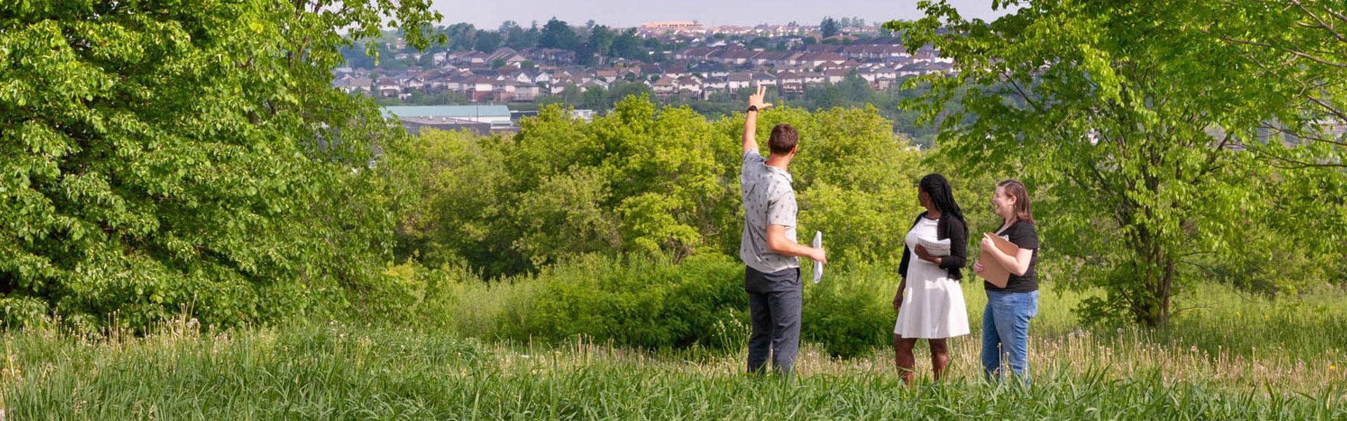 Three students stand in grass and look out at small city in the distance