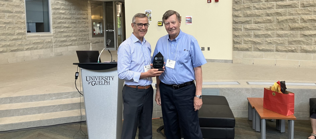 Dr. Ian Dohoo (left) presents Dr. Wayne Martin (right) with a small award made of dark engraved glass to commemorate his being honoured with the CAVEPM Lifetime Leadership Award. The two stand in front of a stage in a brick-walled atrium at the CAVEPM annual meeting. 