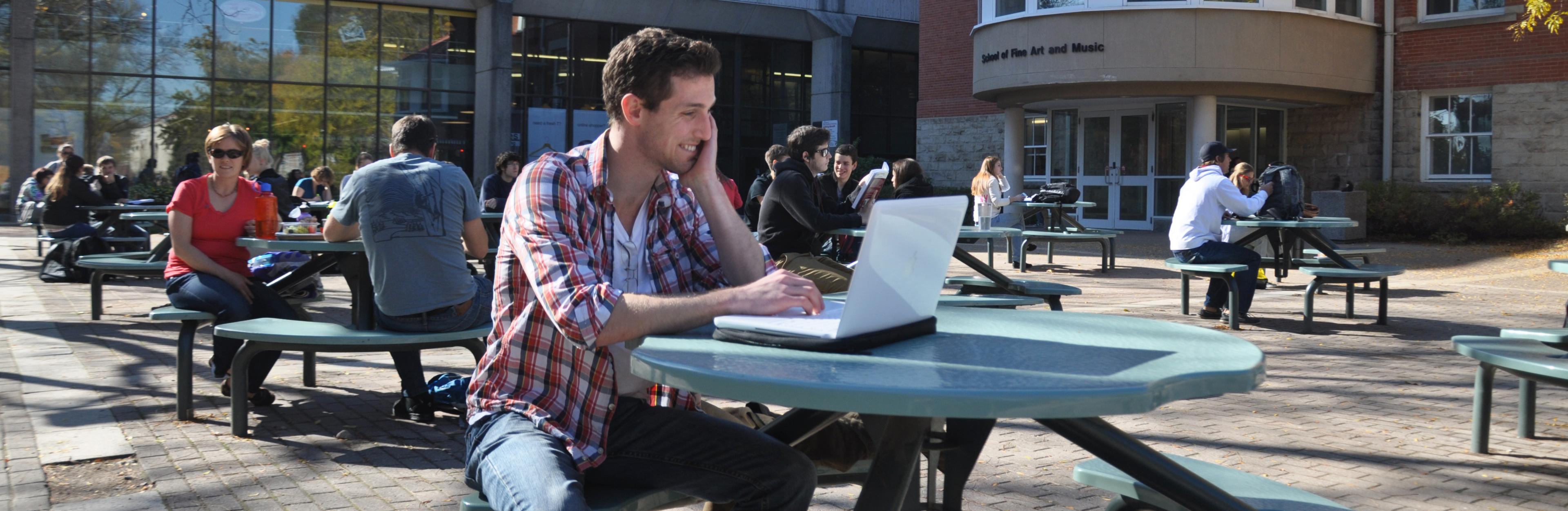 Picture of student sitting at a picnic table outside while working on a laptop. 