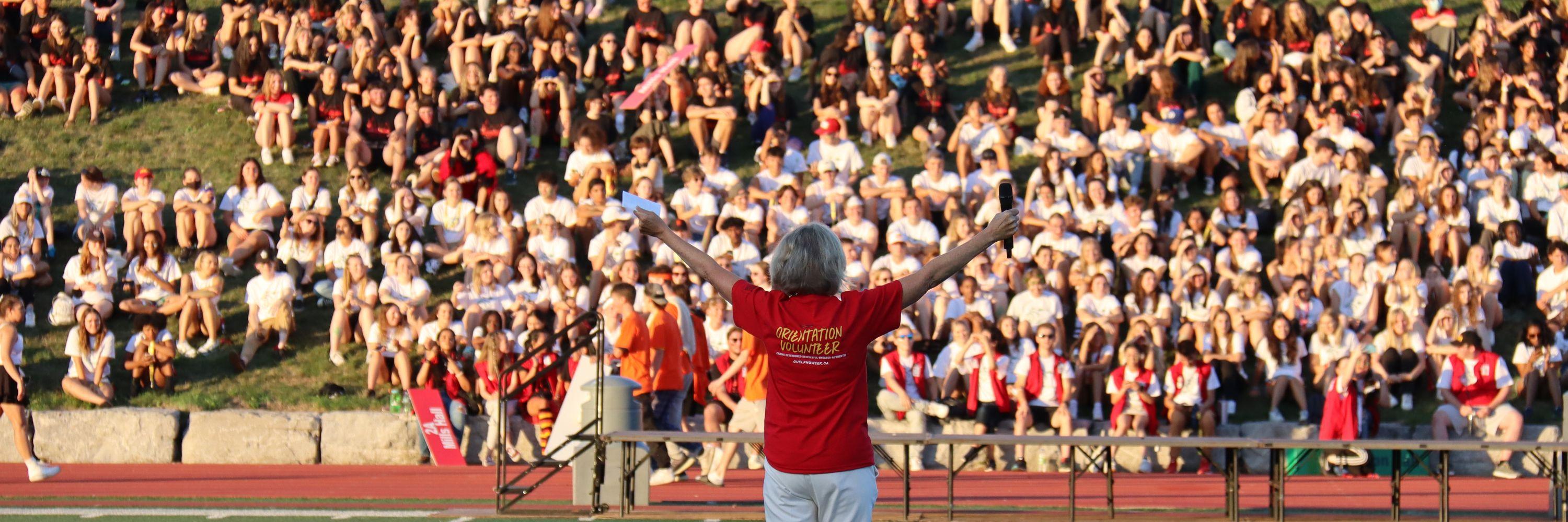 Photograph of Charlotte Yates with arms outstretched, addressing a large crowd of students gathered on a hill.