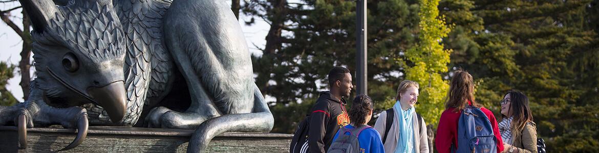 Photo of a group of five students talking to each other in front of the Gryphon statue.