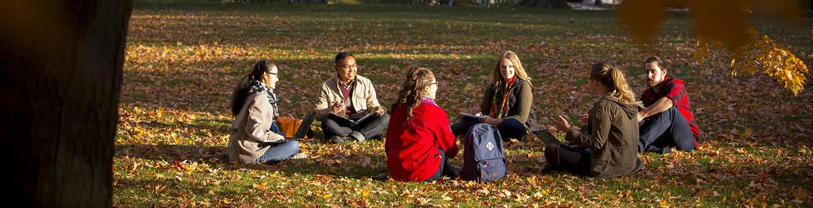 Photo of a group of six students sitting on Johnston Green in the fall, leaves all around them.