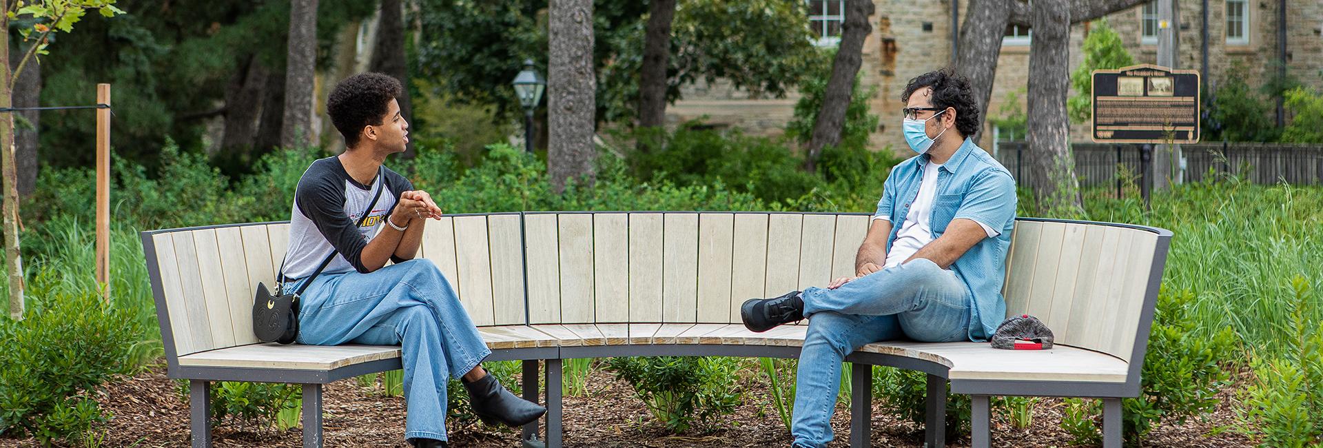 two University of Guelph students sitting on a bench at a distance from each other, chatting.