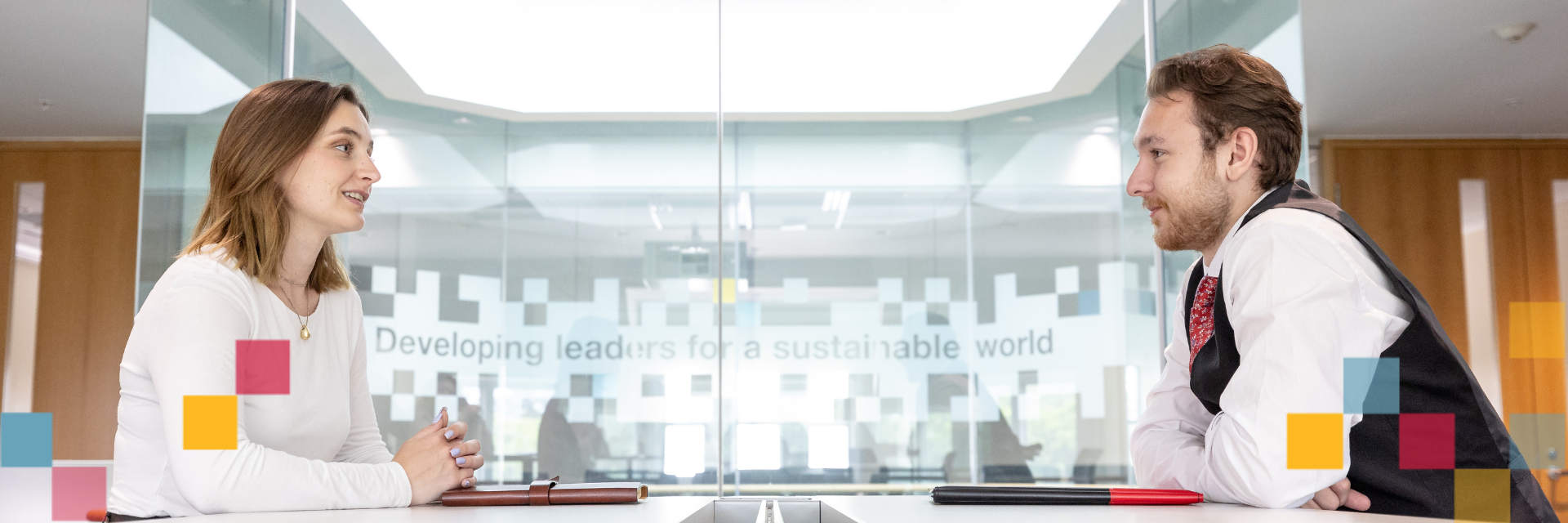 Two people sitting across from each other talking with windows in the background.