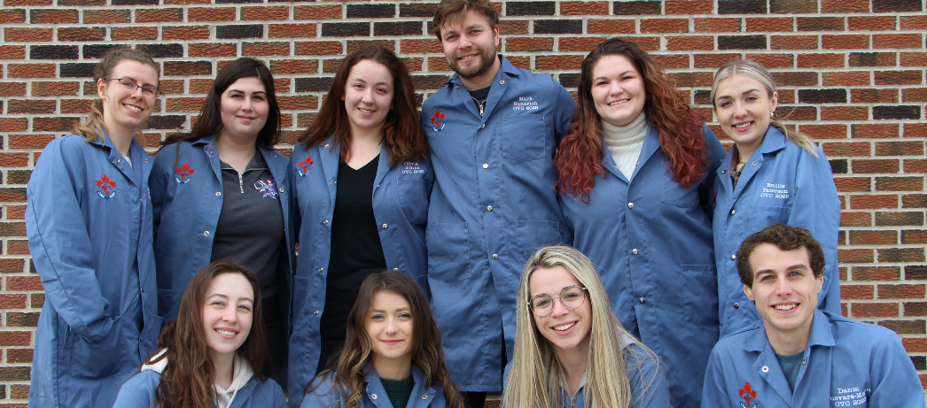 A group of student veterinarians gather in front of a brick wall wearing blue clinical coats.
