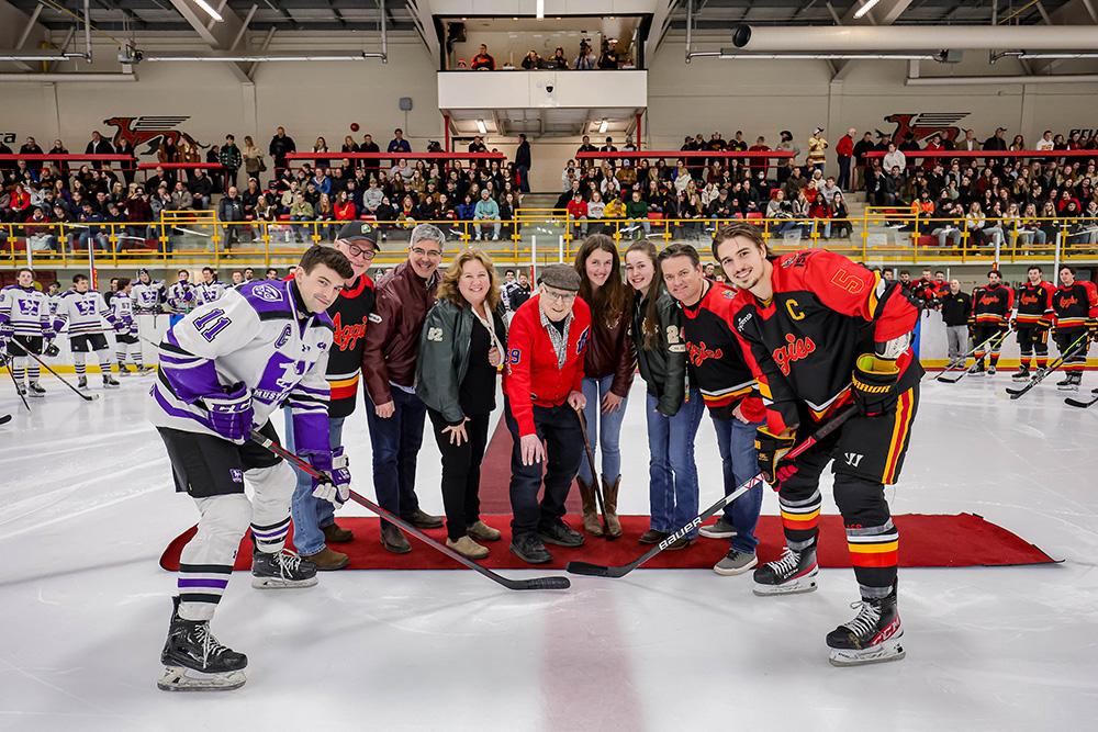 A group of people on a red carpet drop the puck in front of two hockey players
