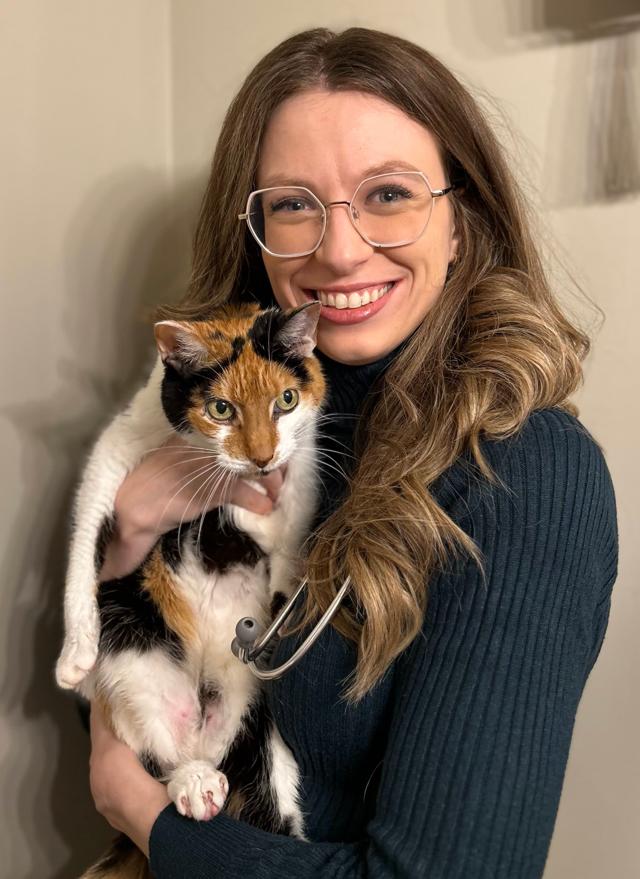 A woman with long hair and glasses smiling and holding a white, brown and black cat