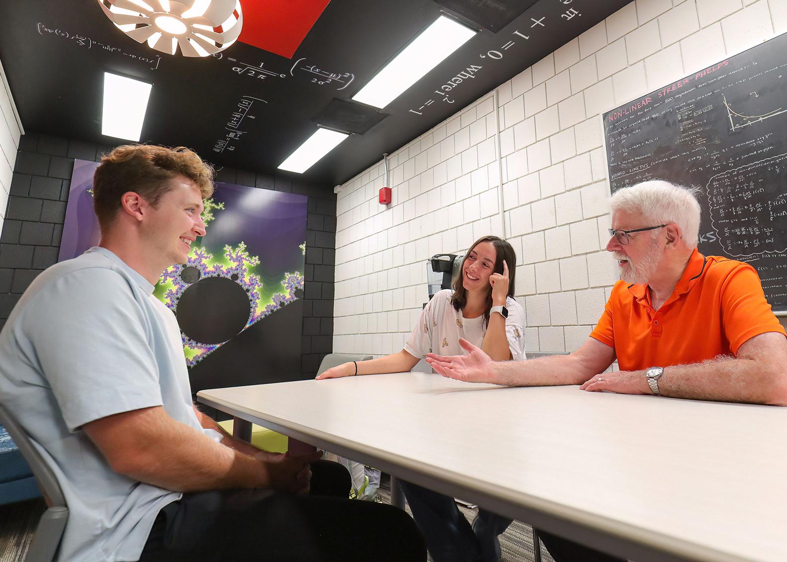 two students and a professor sit at a table and discuss mathematics