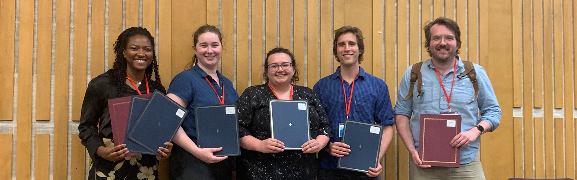 five award winning students standing in a row, in a lecture hall holding award folders