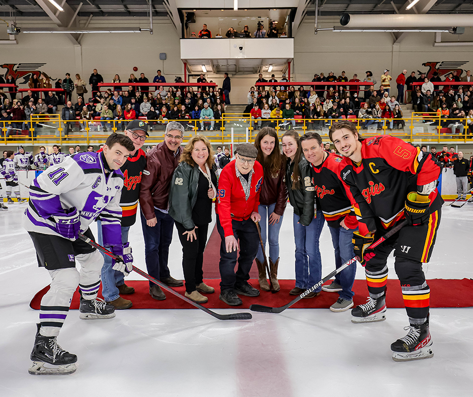 A group of people on a red carpet drop the puck in front of two hockey players