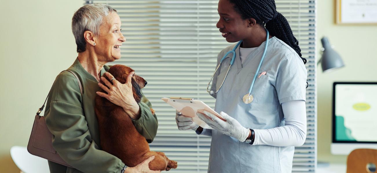 Two people standing in a veterinary office talking. One person is holding a small brown dog.