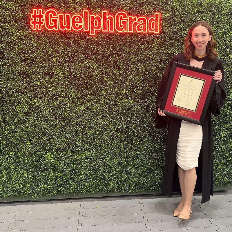 Recent grad standing holding their diploma in front of a #GuelphGrad neon sign.