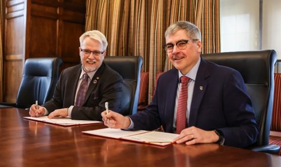 Two men sit side by side, each signing a copy of the agreement and smiling at the camera