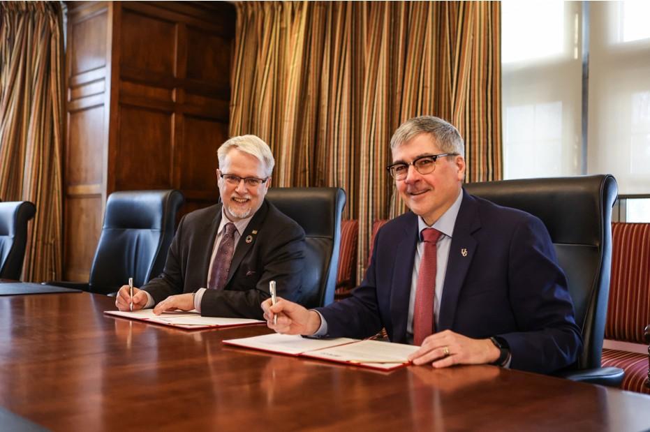 Two men sit side by side, each signing a copy of the agreement and smiling at the camera