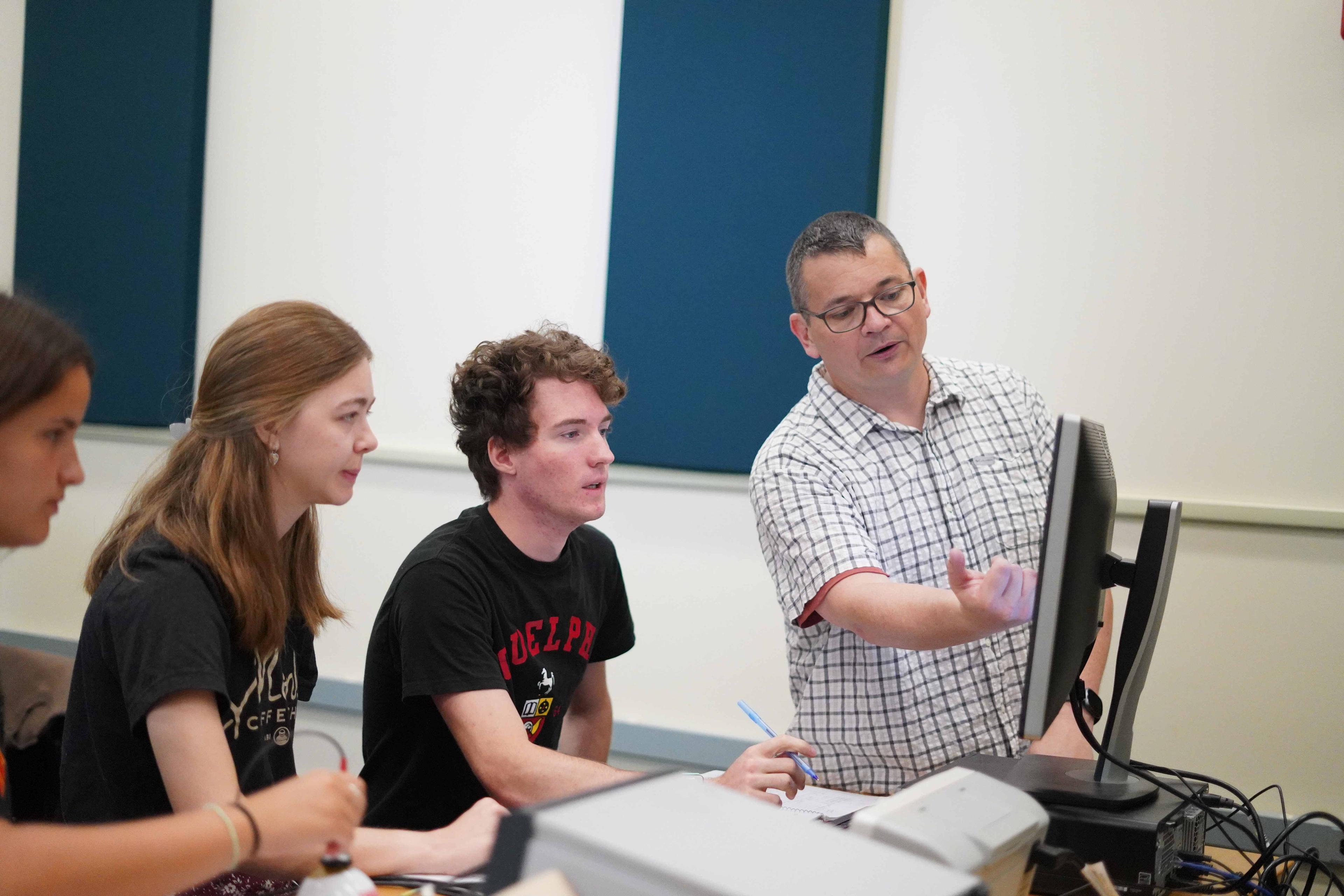 A professor is pointing to a computer screen as he explains to students.