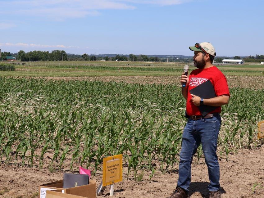Correndo is presenting from a corn field, with plants about knee-height, and speaks into a microphone
