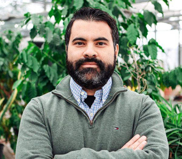 Dr. Adrian Correndo stands in front a leafy plant with arms crossed