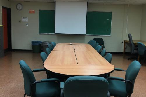 A white walled room containing a long table with several chairs and a projector screen