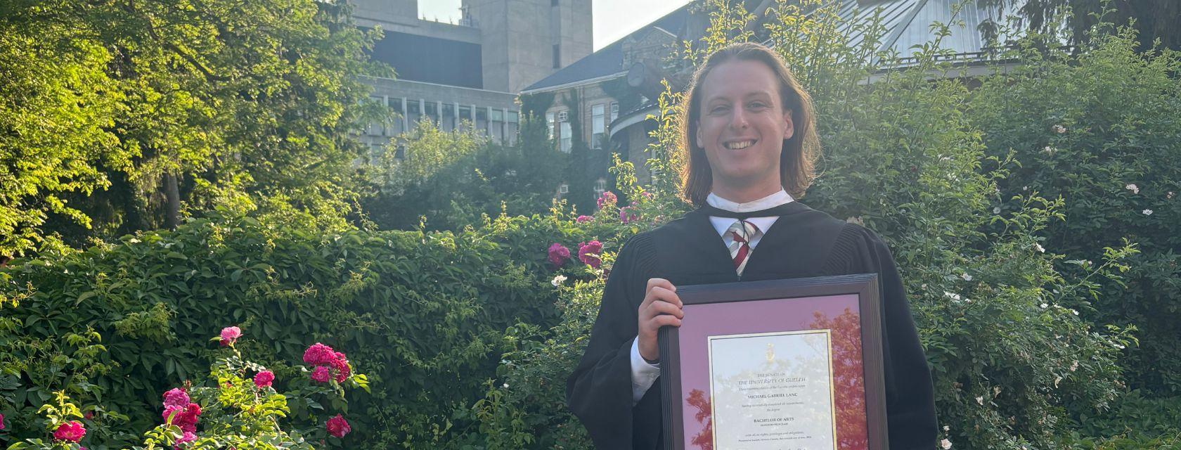 Guelph Grad Michael Lanc poses with his diploma in U of G's commorative gardens.