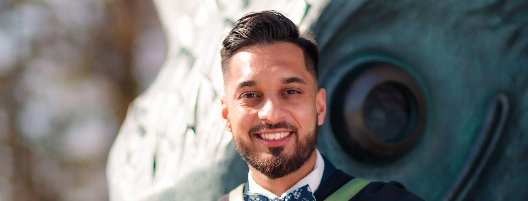 Engineering Grad Abhinav Chatterjee poses at his convocation wearing graduation attire while sitting in front of  a large baby gryphon statue.