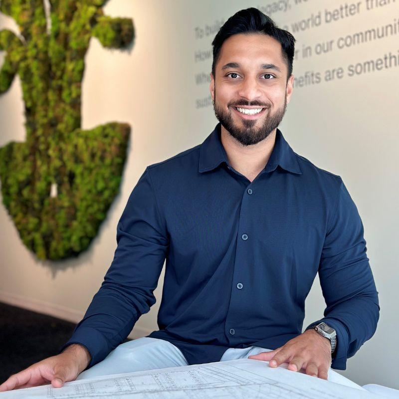Engineering Grad Abhinav Chatterjee sits a desk with blue prints in front of him with a large white wall in the background displaying text and a mossed green wall. 