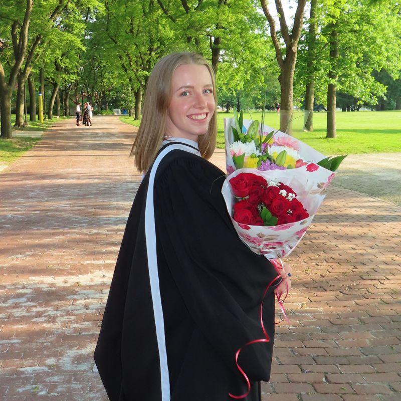 Guelph grad Alexandra Crowley standing outside on a cobblestone path in front of green trees while holding flowers and wearing her graduation attire. 