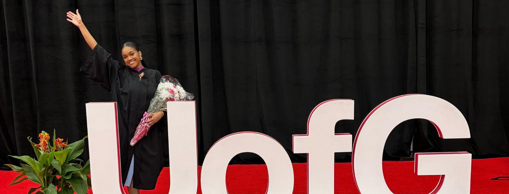 Guelph Grad Marika Bowrin poses at her convocation wearing graduation attire on a red carpet while posing in front of a large letters displaying "UofG".  