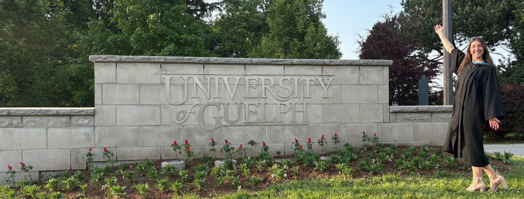 Guelph Grad Paulina Morelli stands in front of a stonewall sign that says University of Guelph while she is wearing her graduation attire. 