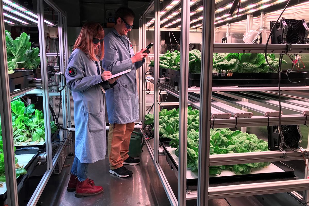 Two students writing on a clipboard in front of plants in a greenhouse