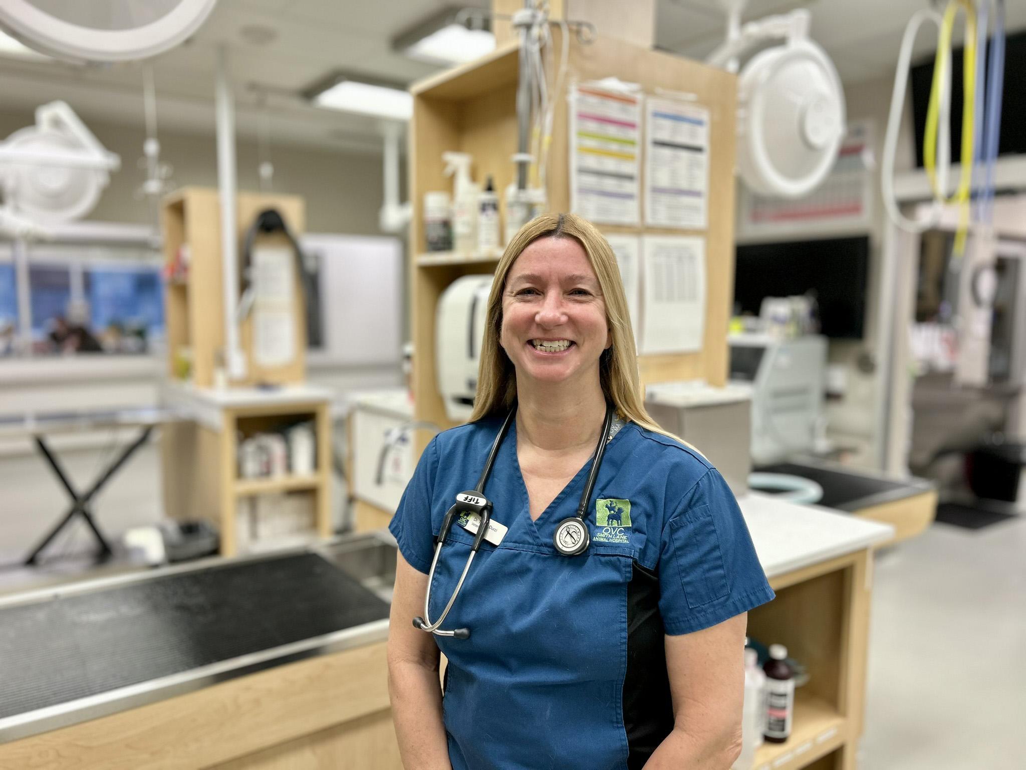 A woman wearing blue hospital scrubs and a stethoscope around her neck smiles at the camera