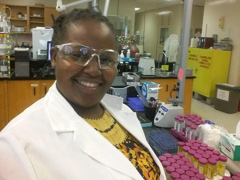 Beatrice Wausi Gedion poses in the lab with samples wearing a lab coat and safety glasses.