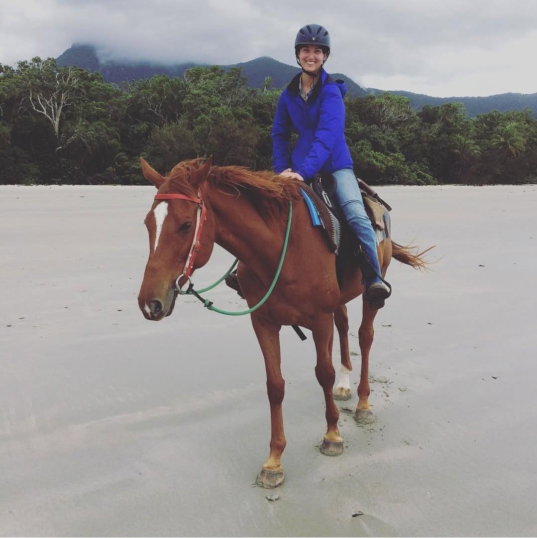 Woman on a horse on a beach with trees and mountains in the background.