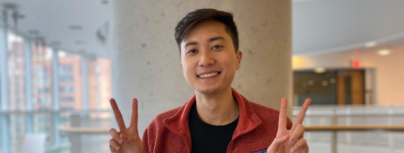 Computer Science Grad Dennis Kao smiling indoors and holding both hands up with the index and middle fingers extended upward in a V-shape while the other fingers are folded down, back of the hand facing forward creating two peace signs .