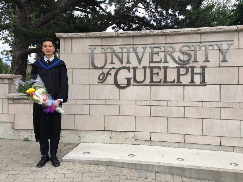 Guelph Grad Dennis Kao posing with flowers outside the U of G sign at his convocation.