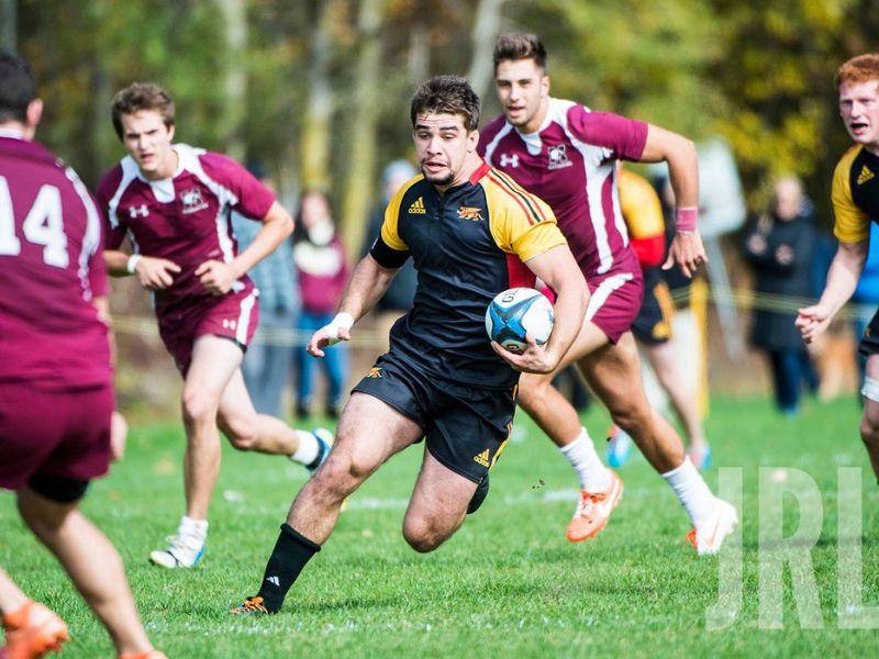 Guelph Grad Jeremy Wright holding a rugby ball while running on the rugby field while multiple players from the opposing team surround him.