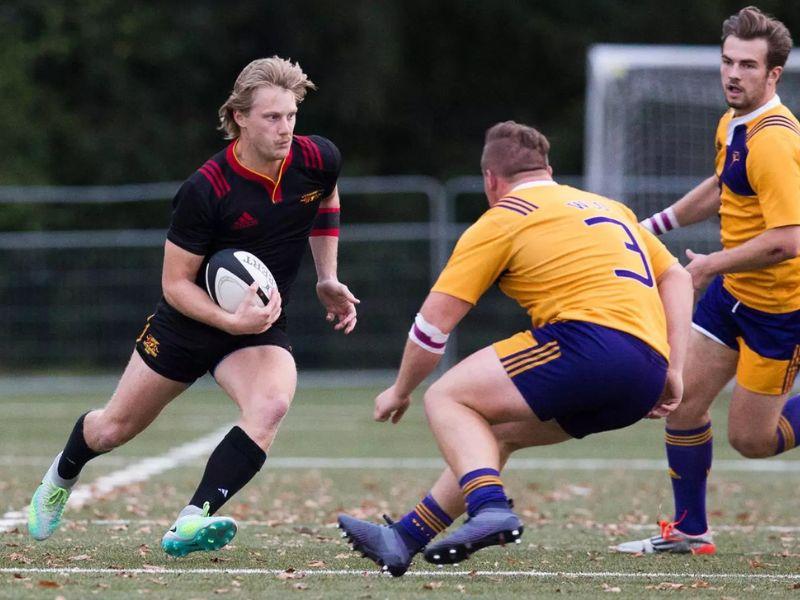 Guelph Grad Jordan Hofstra holding a rugby ball while avoiding two players on the opposing team who are wearing yellow jerseys.