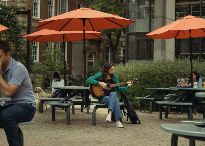 Student playing guitar at a table in a crowded outdoor study space
