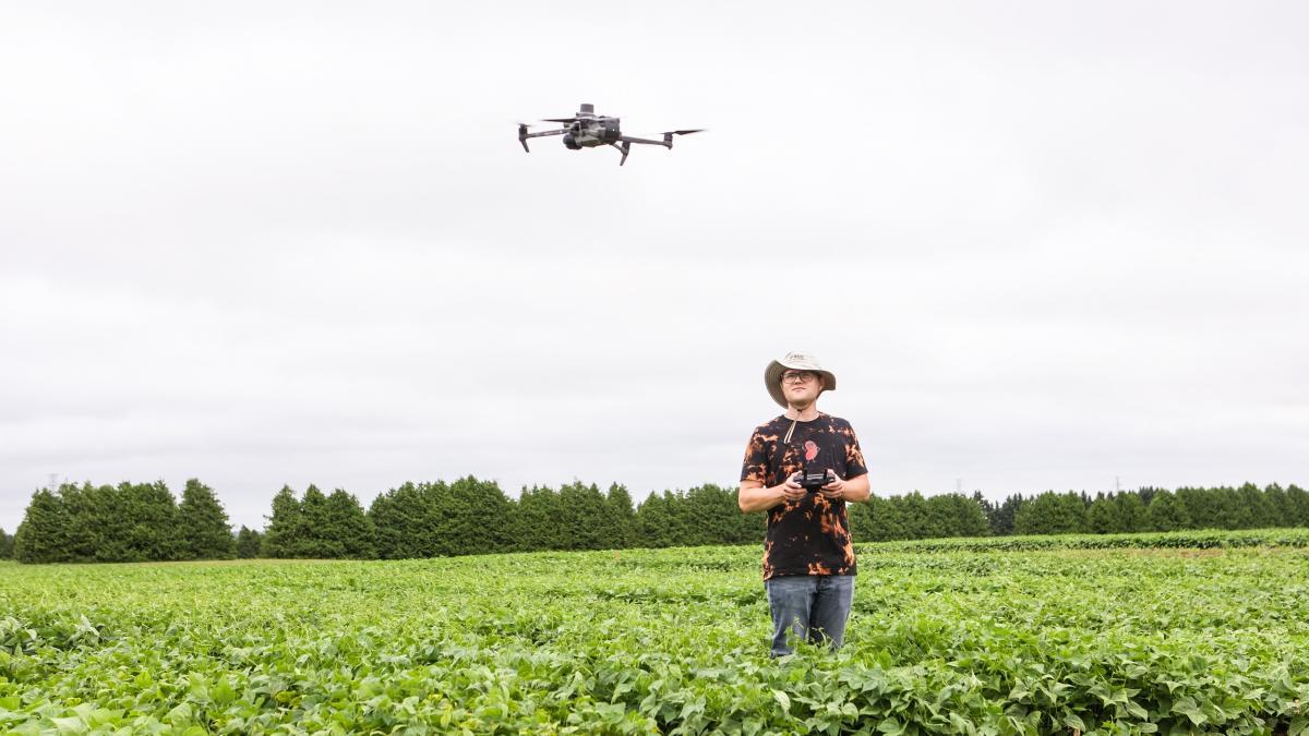 Student standing in a field holding a drone controller while a drone hovers nearby