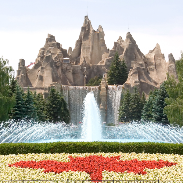 Large fountain in front of rugged, mountain-like rock structures, with flower beds and trees in a landscaped theme park setting.