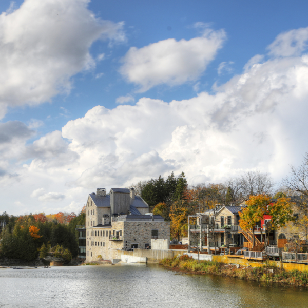 Stone mill building beside a calm river, with autumn trees and a partly cloudy blue sky overhead.