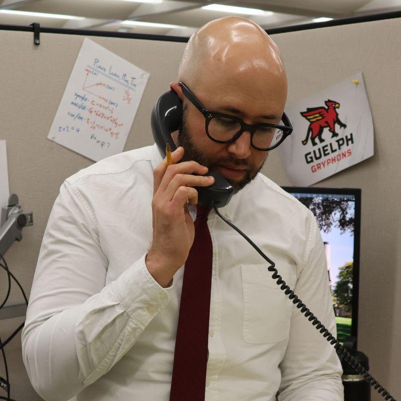 The host of faculty in 15 Ryan holds a phone to his ear while wearing a button up shirt with a tie